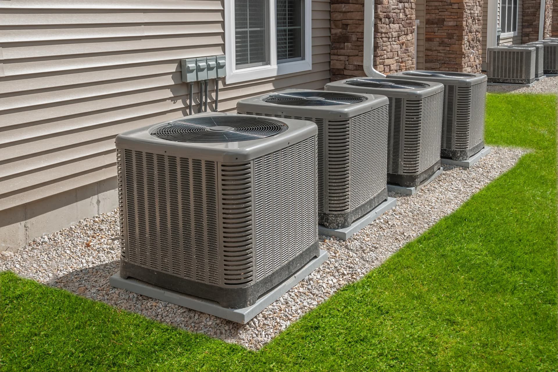 Multiple outdoor air conditioning units installed along the side of a building with light brown siding and stone facade.